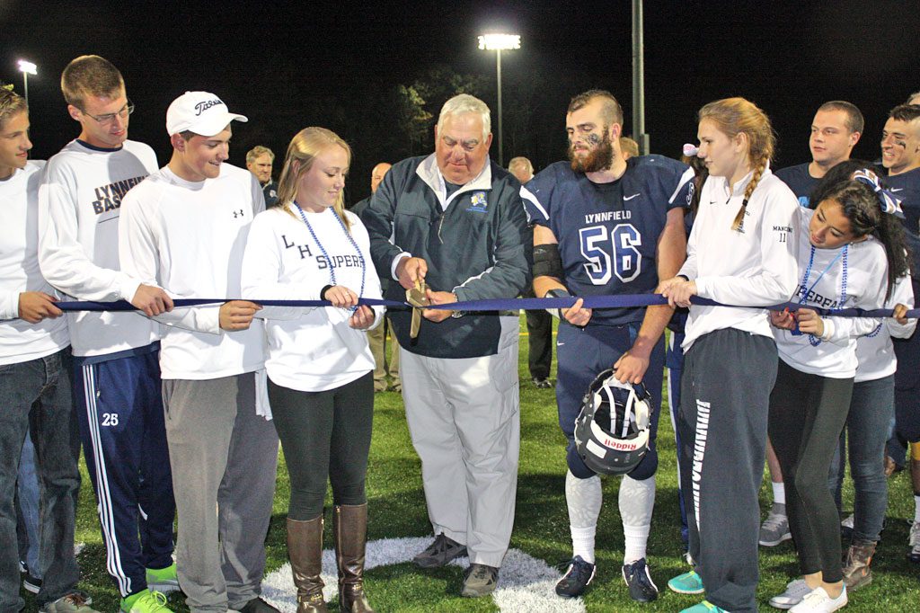 FIELDS COMMITTEE CHAIRMAN Arthur Bourque cuts the ribbon for the $6.5 million Lynnfield High School fields complex while being assisted by LHS captains and student-athletes during the grand opening ceremony on Friday, Oct. 10. The halftime ceremony took place during the football team’s home opener against Georgetown.